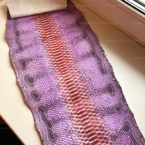May include: A flat, rectangular piece of dyed snakeskin leather featuring a vibrant purple and pink gradient. The skin displays a distinct, textured scale pattern with a prominent, lighter-colored central ridge running vertically down the length.
