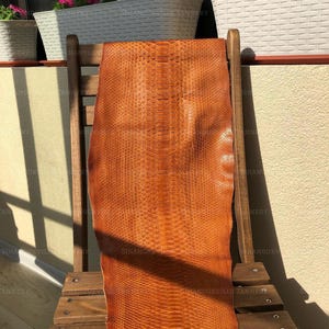 Puede incluir: Una pieza larga y rectangular de piel de serpiente natural en un vibrante color naranja bronceado. El material presenta un patrón de escamas simétrico y distintivo que recorre verticalmente el centro, con una superficie texturizada.