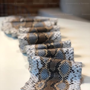 May include: A series of rolled pieces of snakeskin leather featuring a repeating diamond pattern in shades of white, grey, and brown. The text Sihanoukville Tannery is repeated across the image in a watermark style over the textured material.