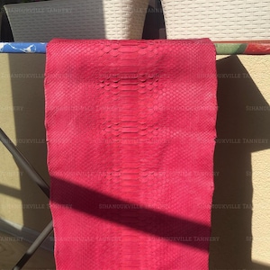 May include: A vibrant hot pink snakeskin leather hide featuring a distinct, repeating scale pattern across the entire surface. The material is draped over a horizontal bar, displaying a uniform texture and a consistent, saturated color throughout.