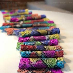 May include: A collection of rolled snakeskin leather pieces featuring a vibrant, multi-colored scale pattern in shades of pink, blue, yellow, green, and red. Each piece displays the distinct, textured diamond-shaped pattern of snakeskin.
