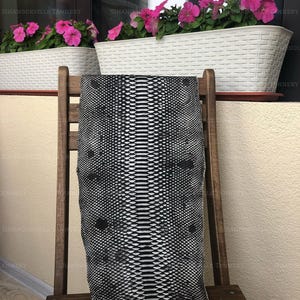 May include: A rectangular piece of black and white snakeskin leather featuring a distinct scale pattern with a central spine line. The hide is draped over a wooden chair, displaying a high-contrast monochromatic texture throughout the surface.