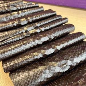 Può includere: Una pila di pezzi di pelle di serpente piegati in bronzo metallizzato e marrone scuro. Il materiale testurizzato presenta un motivo a scaglie ripetitivo con una lucentezza riflettente. Il testo SIHANOUKVILLE TANNERY è visibile come filigrana.
