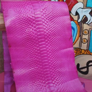 May include: A vibrant hot pink snakeskin leather hide featuring a distinct, symmetrical scale pattern running down the center. The material has a glossy, textured surface with small, uniform scales that transition into larger, elongated scales.