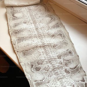 May include: A long, flat piece of natural snakeskin leather featuring a repeating scale pattern in shades of off-white, cream, and light gray. The hide displays a distinct central spine line with symmetrical, diamond-shaped markings extending outward.