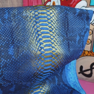 May include: A piece of textured material featuring a snakeskin pattern in shades of deep blue and black. A prominent central stripe runs vertically down the middle, displaying a metallic gold finish on the raised, overlapping scale-like segments.