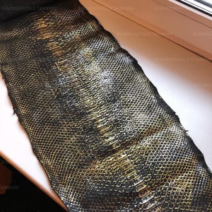 May include: A long, rectangular piece of snakeskin leather featuring a distinct scale pattern. The surface is finished in a deep black color with a metallic gold wash applied to the raised edges of the scales, creating a high-contrast appearance.