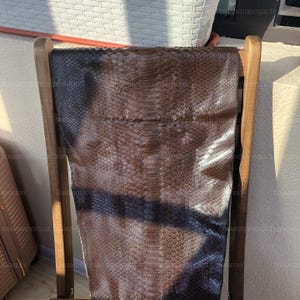May include: A large, rectangular piece of dark brown snakeskin leather with a distinct, repeating scale pattern. The hide features a natural gradient of deep brown tones, with a prominent central ridge running vertically down the length of the skin.