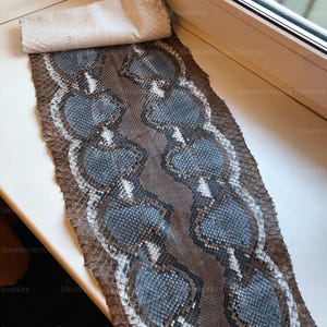 May include: A flat, rectangular piece of snakeskin leather featuring a repeating pattern of blue-grey, heart-shaped scales bordered by dark brown and white edges. The top section is rolled over, revealing a plain, light-colored underside.
