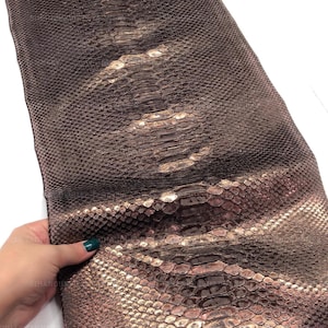 May include: A long, rectangular piece of dark brown snakeskin leather featuring a distinct, repeating scale pattern. The surface displays a metallic, copper-toned sheen across the central scales, transitioning into a deep, matte brown texture along the edges.