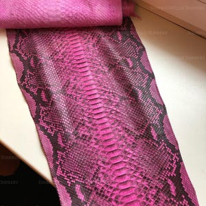 May include: A piece of genuine snakeskin leather dyed in a vibrant hot pink color with a natural black scale pattern. The hide features a distinct central row of large, elongated scales flanked by smaller, textured scales throughout the surface.