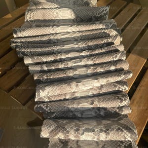 Puede incluir: Una pila de varias pieles de cuero de serpiente grises y blancas, cada una con un patrón de escamas natural distintivo. La superficie del material muestra una apariencia reptiliana texturizada con varios tonos de gris claro, carbón y blanco.
