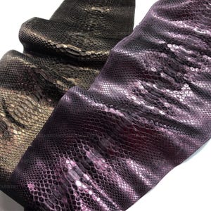 May include: Two pieces of textured snakeskin leather are displayed side by side. One piece is a deep, metallic purple with a glossy finish, while the other is a dark, muted charcoal gray with a subtle sheen, both showing natural scale patterns.