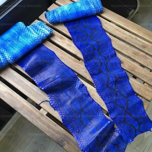 May include: Two long, rectangular strips of blue snakeskin leather featuring a distinct, repeating diamond-shaped scale pattern in deep blue and black. The material has a glossy finish and is displayed in a rolled and flat configuration.