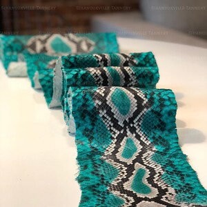 May include: A long, folded strip of snakeskin leather featuring a vibrant turquoise, black, and white diamond-shaped scale pattern. The material has a textured, reptilian surface with a repeating geometric design and the text SIHANOUKVILLE TANNERY.