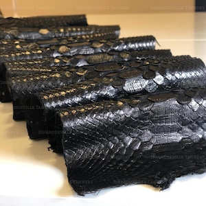 Puede incluir: Una serie de piezas de piel de serpiente negra dispuestas en fila. Cada pieza presenta un patrón de escamas superpuestas distintivo con un acabado brillante. El texto SIHANOUKVILLE TANNERY es visible en un patrón repetido en la imagen.