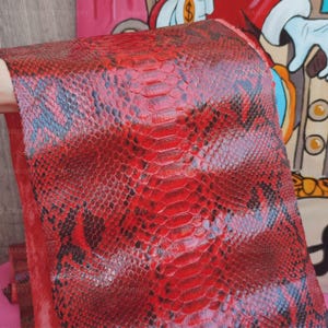 May include: A piece of red and black snakeskin leather featuring a distinct, repeating scale pattern. The material displays a vibrant crimson hue with darker, charcoal-colored markings integrated into the texture of the individual scales.