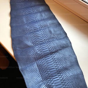 May include: A long, rectangular piece of blue snakeskin leather featuring a distinct, repeating scale pattern across its entire surface. The material displays a consistent deep blue hue with a textured finish and the text SIHANOUKVILLE TANNERY.