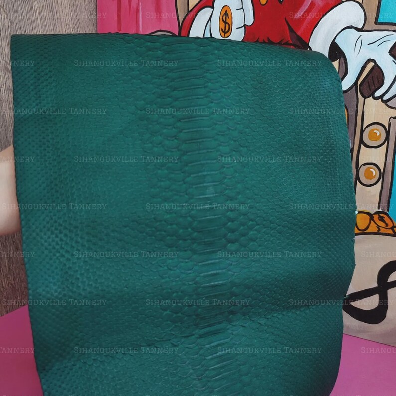 May include: A rectangular piece of dark green snakeskin leather featuring a distinct central row of larger, elongated scales flanked by smaller, textured scales. The surface is imprinted with the repeating text Sihanoukville Tannery.