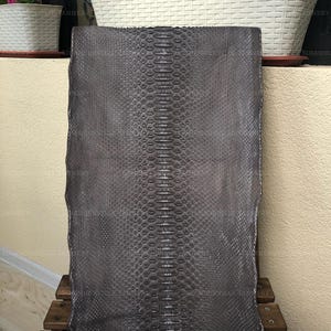 May include: A rectangular piece of dark gray snakeskin leather featuring a prominent central row of large, symmetrical scales flanked by smaller, fine-textured scales on both sides. The hide displays a consistent matte finish and natural pattern.