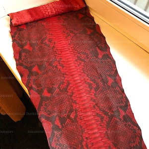 May include: A flat, elongated piece of genuine snakeskin leather dyed in a vibrant red and dark black color scheme. The material features a distinct, natural scale pattern throughout, with a rolled top edge and a visible watermark text overlay.