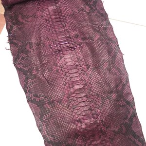 May include: A deep burgundy and black snakeskin hide featuring a prominent central row of large, elongated scales flanked by smaller, textured scales. The surface displays a natural, repeating pattern with subtle variations in color depth and scale size.
