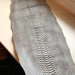 May include: A long, rectangular piece of grey and white snakeskin leather featuring a distinct, symmetrical scale pattern running down the center. The surface displays a natural, textured appearance with the text SIHANOUKVILLE TANNERY visible.