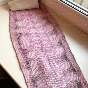 May include: A long, flat piece of snakeskin leather dyed in shades of pink and grey. The hide features a distinct, symmetrical scale pattern running down the centre, with a white paper towel roll placed at the top edge of the textured material.