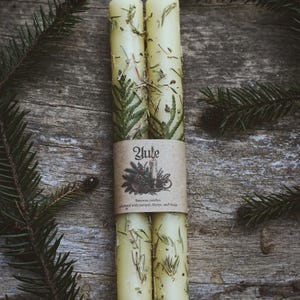 May include: Two pale yellow beeswax taper candles embedded with dried juniper, thyme, and thuja sprigs. A brown paper band wraps around the center, featuring the text Yule, an illustration of a candle, and the words Beeswax candles adorned with juniper, thyme, and thuja.