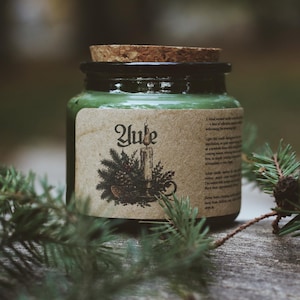 May include: A green candle in a glass jar with a cork lid. The label features the text Yule, an illustration of a candle with pine branches, and printed instructions. The jar is surrounded by green pine needles on a textured surface.