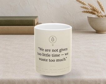 Puede incluir: Una taza de cerámica de color crema con un icono circular negro que muestra un pequeño diseño de hoja sobre el número romano II. El texto dice: "We are not given too little time - we waste too much." La taza tiene una forma cilíndrica.