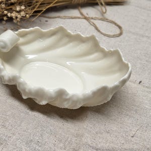 May include: A cream-coloured ceramic dish shaped like a curled leaf with a glossy finish. The edges of the dish feature intricate, ruffled detailing that mimics the natural texture of foliage, with a prominent scroll at one end of the rim.