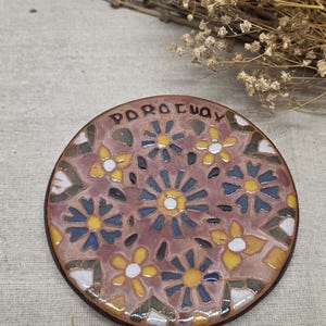 Könnte beinhalten: Ein runder Keramikuntersetzer mit einem floralen Mosaikmuster in den Farben Mauve, Gelb, Blau und Weiß. Das Wort POROLUA ist in schwarzen Buchstaben am oberen Rand eingraviert. Die Oberfläche hat ein glänzendes Finish mit sichtbaren Fugen.