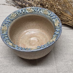 Op de afbeelding: Een ronde keramische kom met een gespikkelde beige binnenkant en een gestructureerde, blauwachtige rand met verhoogde, lichtgekleurde decoratieve markeringen. De kom heeft een gladde, glanzende afwerking aan de binnenkant en een ruwere buitenkant.