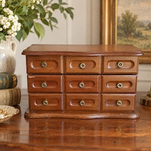 May include: A wooden storage chest featuring nine rectangular drawers arranged in three rows of three. Each drawer has a recessed oval panel and a small, round, gold-toned metal knob with a textured edge. The chest has a smooth, polished finish.
