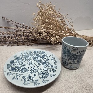 May include: A white ceramic plate and a matching cup, both decorated with a detailed dark blue line drawing of a coastal village scene featuring houses, trees, and sailing ships on the water, presented as a coordinated set of tableware.