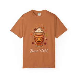 May include: A burnt orange short-sleeve t-shirt featuring a graphic of a coffee cup with whipped cream, a pumpkin, and cinnamon sticks. The cup has a jack-o'-lantern face and text reading "PUMPKIN SPICE & SPOOKY VIBES EST. OCTOBER" and "Basic Witch".