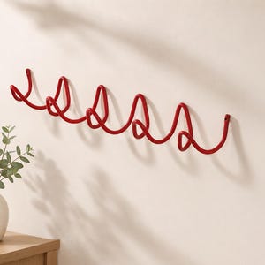May include: A wall-mounted coat rack made of a single continuous piece of red metal bent into a series of five interconnected, swirling loops. The structure features mounting points at both ends and provides multiple hooks for hanging items.