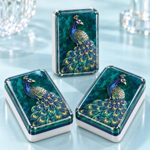 May include: Three rectangular boxes with white sides and a dark teal marbled top surface. Each lid features a raised, detailed metal peacock design in shades of blue, green, and gold, with a prominent tail display and a small crest on the head.