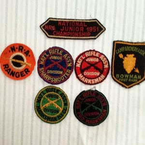 May include: A collection of seven embroidered fabric patches featuring various designs and text including National Rifle Association Junior Championship 1951, NRA Ranger, Sharpshooter, Marksman, Pro-Marksman, and Camp Archery Association Bowman First Rank.