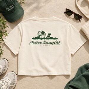 May include: A cream-colored short-sleeve t-shirt featuring a dark green graphic on the back. The design includes a globe, a tower, and the text "Mediocre Running Club NEW YORK" in a stylized script font with horizontal lines.