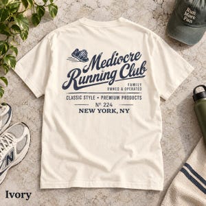 May include: An ivory short-sleeve t-shirt featuring a dark blue graphic on the back that reads Mediocre Running Club, Family Owned & Operated, Classic Style, Premium Products, No. 224, New York, NY, with a small illustration of a running shoe.