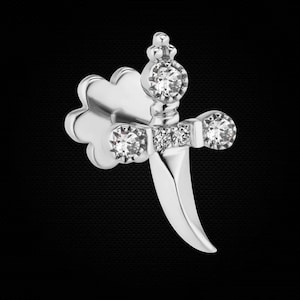 May include: A silver-toned stud earring shaped like a dagger with a curved blade. The hilt features a central clear round gemstone, two smaller clear round gemstones on the crossguard, and a small clear round gemstone at the pommel.