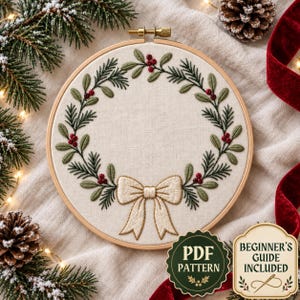 May include: A circular wooden embroidery hoop containing a cream-colored fabric with a hand-stitched wreath design featuring green pine needles, mistletoe leaves, and red berries, finished with a central cream-colored bow at the bottom.
