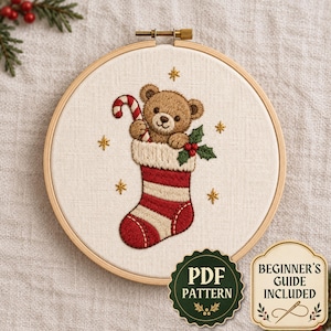 Puede incluir: Un bastidor de bordado circular de madera que contiene un panel de tela con un diseño bordado a mano de un oso de peluche marrón dentro de un calcetín navideño a rayas rojas y blancas. El oso sostiene un bastón de caramelo, rodeado de estrellas doradas y hojas de acebo.
