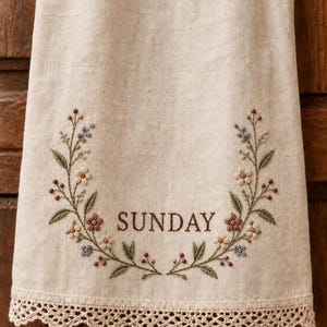 May include: A beige linen tea towel featuring an embroidered floral wreath in shades of pink, blue, and green. The word SUNDAY is stitched in the centre in brown thread. The bottom edge is finished with a delicate, scalloped cream lace trim.