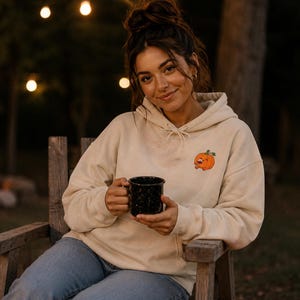 May include: A cream-colored hooded sweatshirt featuring a small, embroidered orange pumpkin graphic on the left chest, paired with blue denim jeans and a black speckled enamel mug held in both hands.