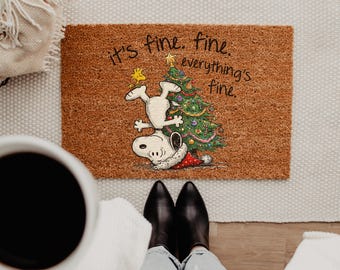 May include: A rectangular brown coir doormat featuring a graphic of Snoopy doing a handstand next to a decorated Christmas tree with Woodstock on top. The text reads, it's fine. fine. everything's fine. in black cursive lettering.