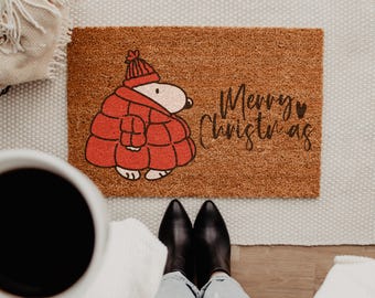May include: A rectangular brown coir doormat featuring a graphic of a white dog wearing a red puffer jacket and a matching red knit hat. The text Merry Christmas is printed in black script on the right side, accompanied by a small black heart.