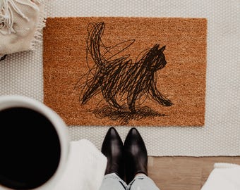 May include: A rectangular brown coir doormat featuring a black graphic illustration of a cat created with a scribbled, chaotic line style. The cat is depicted in profile, walking toward the right side of the mat with its tail held high.